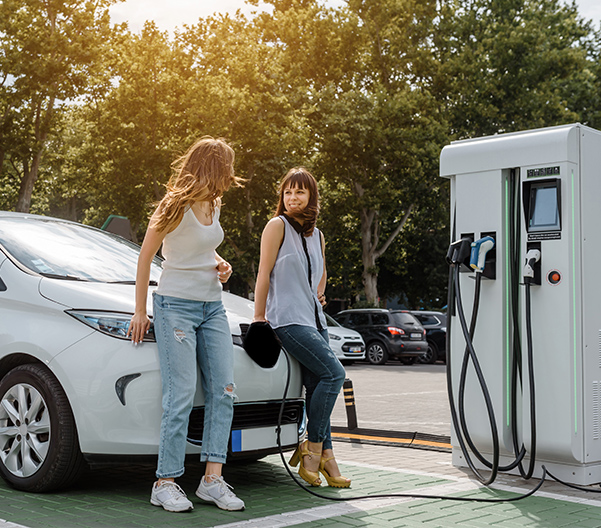 Two young woman resting on hood of car by EV charging station.