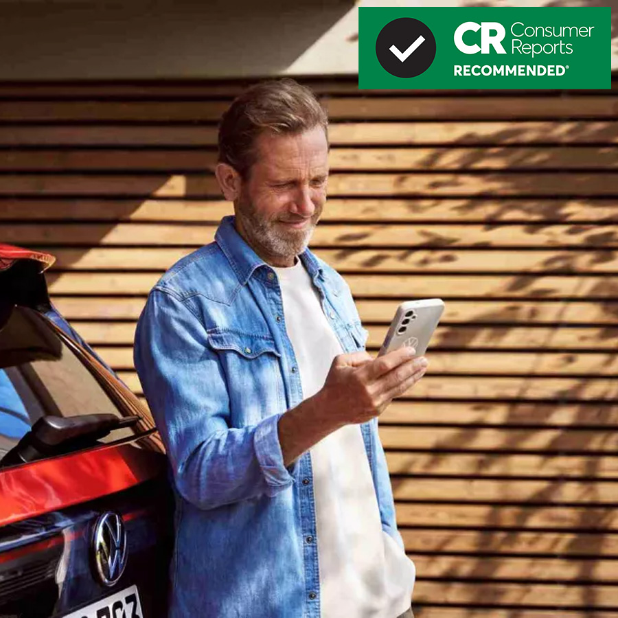 Middle-aged man with a beard in a denim shirt, standing next to a red car while looking at his smartphone